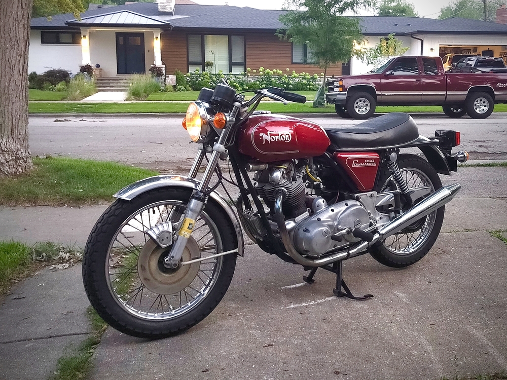 A red 1975 Norton Commando 850 motorcycle with headlight on parked on suburban driveway.