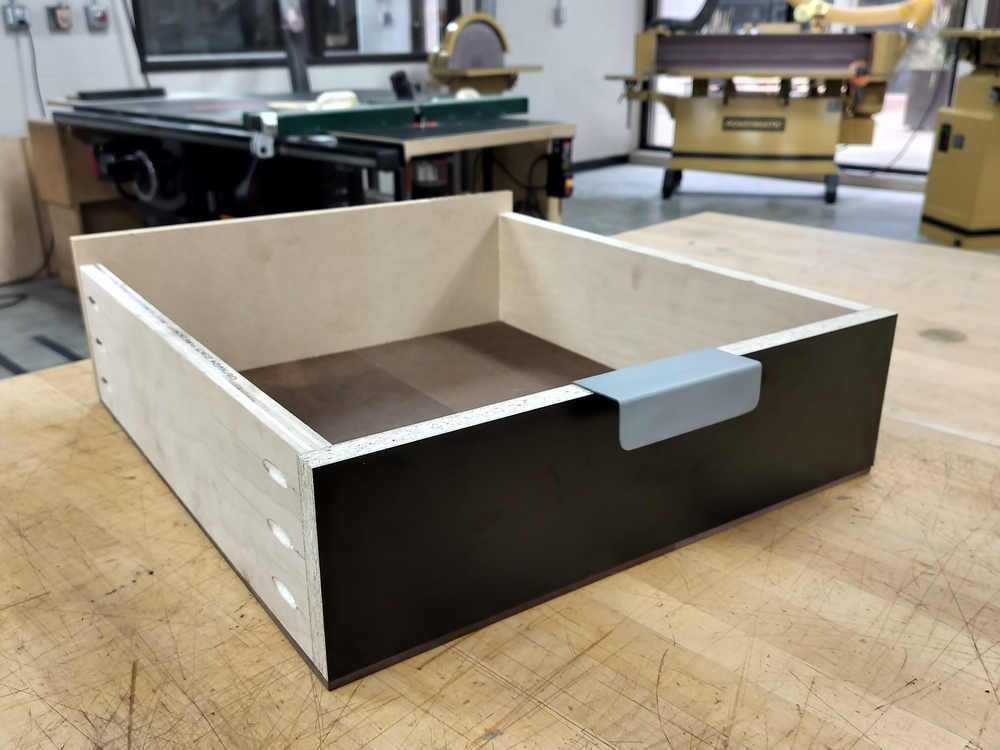 Drawer made from scrap plywood, MDF, and melamine laminated board. Front is made from black finish melamine. The handle is centered towards the top made from bent sheet metal.