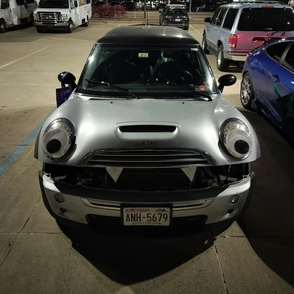 Mini Cooper S R53 with large google eyes on headlights and two cardboard triangle teeth sticking out the bottom of the grill. Hood clamshell is open looking like a mouth about to chomp.