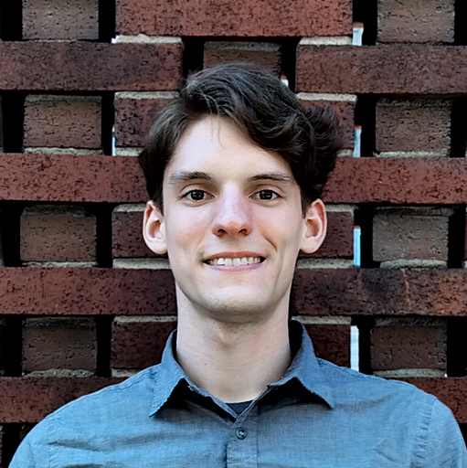 Close up of Alex standing in front of red brick wall wearing a medium gray button down.